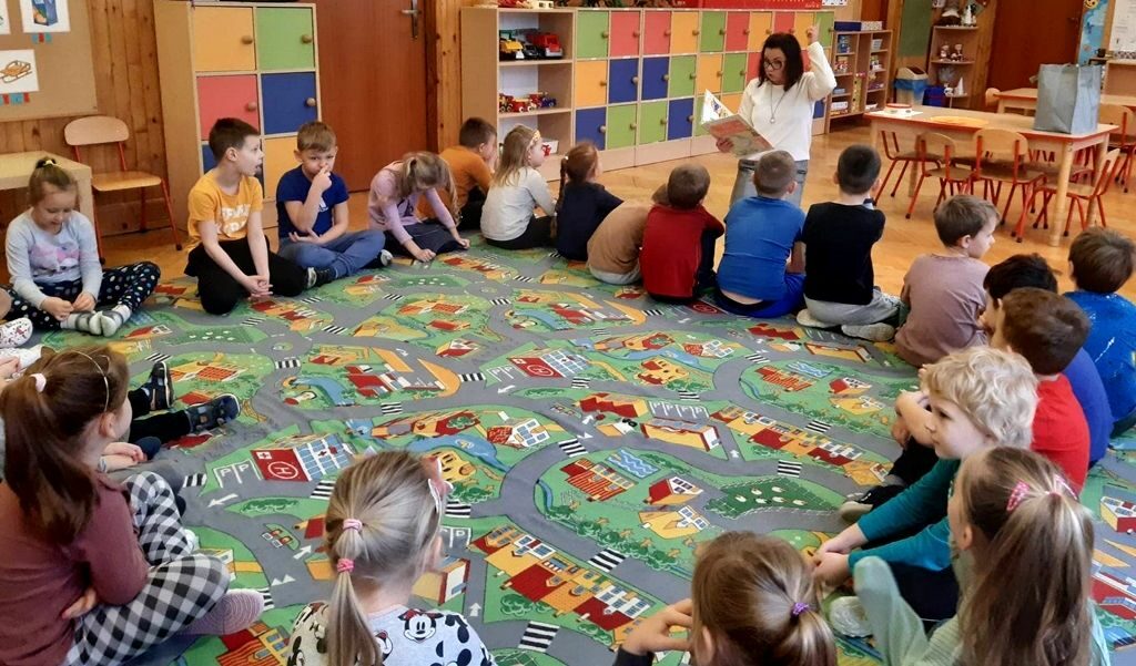 zdjęcie przedstawia kolorową sale przedszkolną, dzieci siedzące na podłodze i słuchające czytającą bibliotekarkę