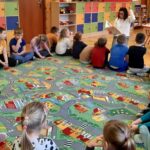 zdjęcie przedstawia kolorową sale przedszkolną, dzieci siedzące na podłodze i słuchające czytającą bibliotekarkę