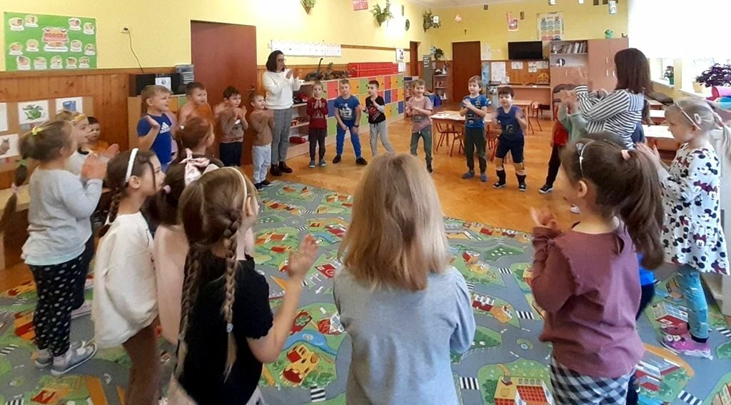 na zdjęciu widać salę przedszkolną i stojące klaskające dzieci.