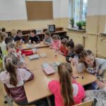 na zdjęciu widać sale szkolną, na środku stoi stół, wokół stołu siedzą dzieci i wykonują prace plastyczną