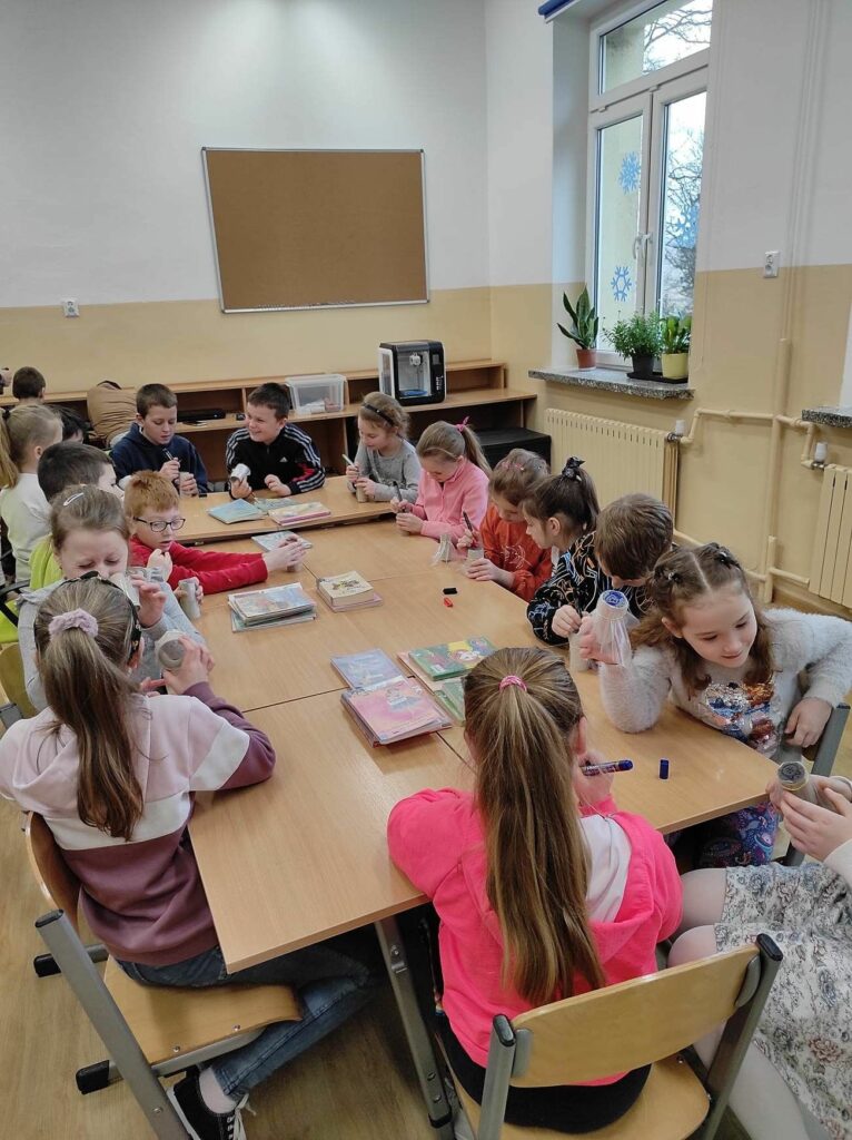 na zdjęciu widać sale szkolną, na środku stoi stół, wokół stołu siedzą dzieci i wykonują prace plastyczną