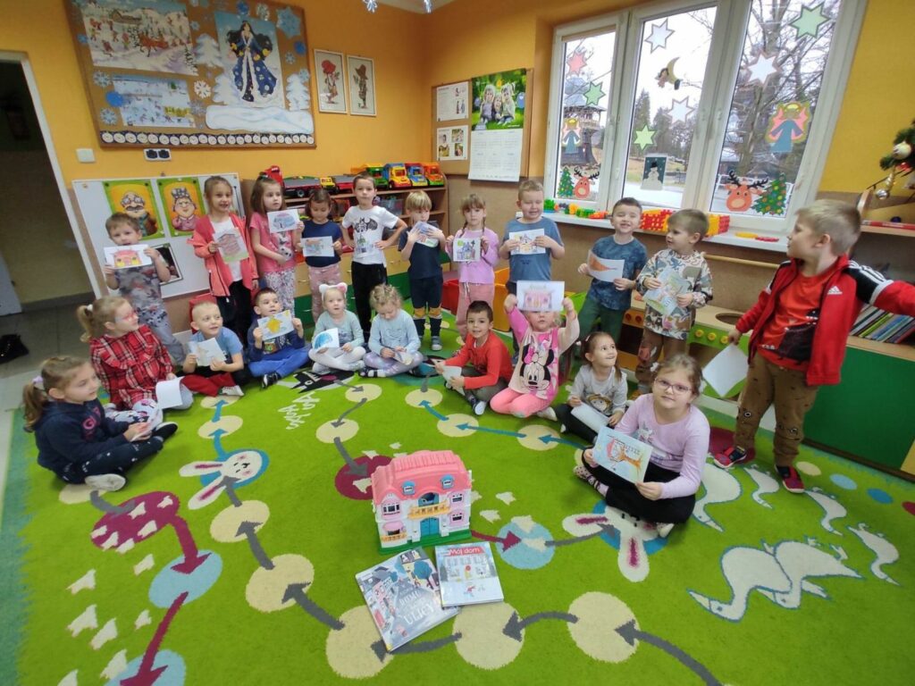 na zdjęciu widać salę przedszkolną, na środku sali stoi domek dla lalek wokół domku siedzą dzieci, prezentują swoje prace plastyczne.
