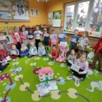 na zdjęciu widać salę przedszkolną, na środku sali stoi domek dla lalek wokół domku siedzą dzieci, prezentują swoje prace plastyczne.