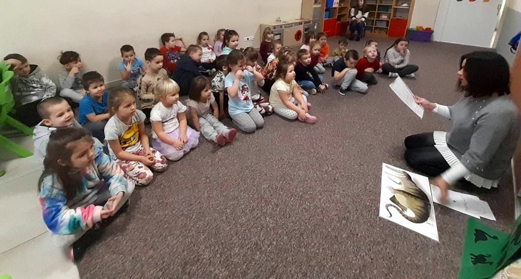 na zdjęciu widać bibliotekarkę czytającą dzieciom .Wszyscy siedzą na podłodze