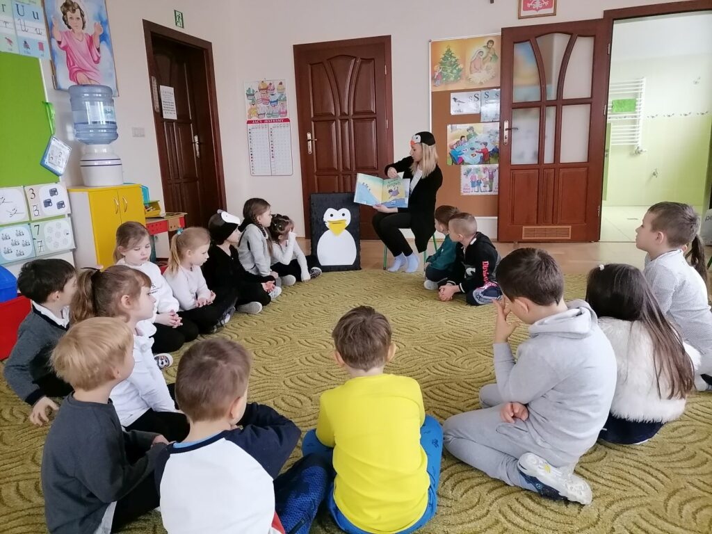 na dywanie w sali w przedszkolu dzieci siedzą w kułeczku na dywanie. Pośród nich siedzi bibliotekarka i pokazuje obrazek w książce, którą wcześniej czytała dzieciom.