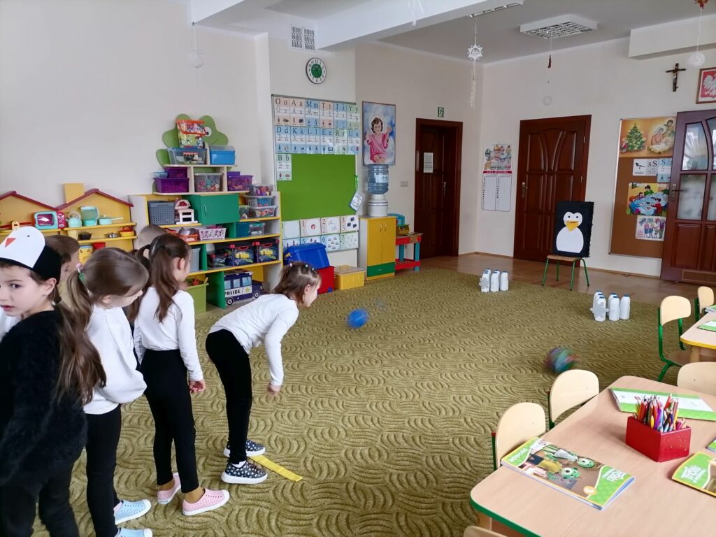 W sali przedszkolnej dzieci rywalizują ze sobą podczas zabawy w rzucie do celu
