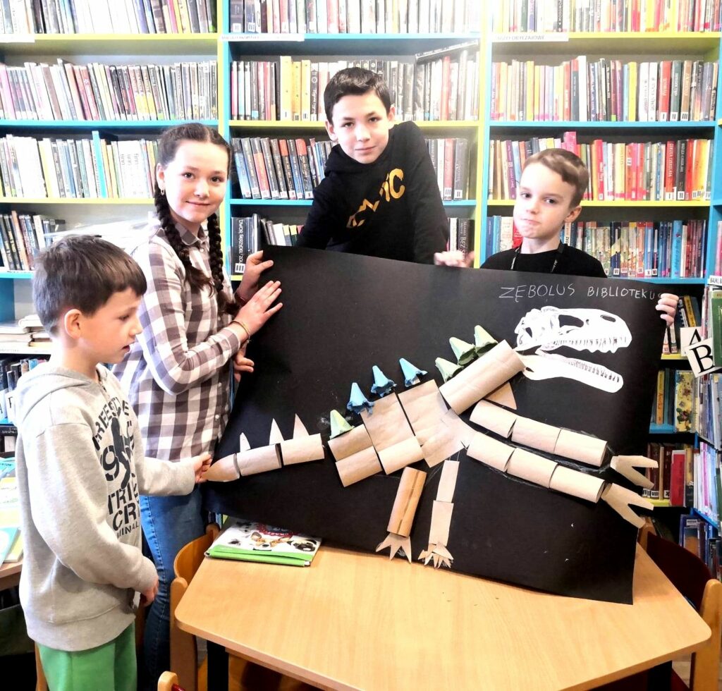na zdjęciu widać dzieci trzymające prace przedstawiającą ogromnego dinozaura