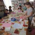 Na zdjęciu widać grupę dzieci siedzących przy stolikach i wykonujących prace plastyczne. Zajęcia odbywają się w bibliotece. W tle regały z książkami.
