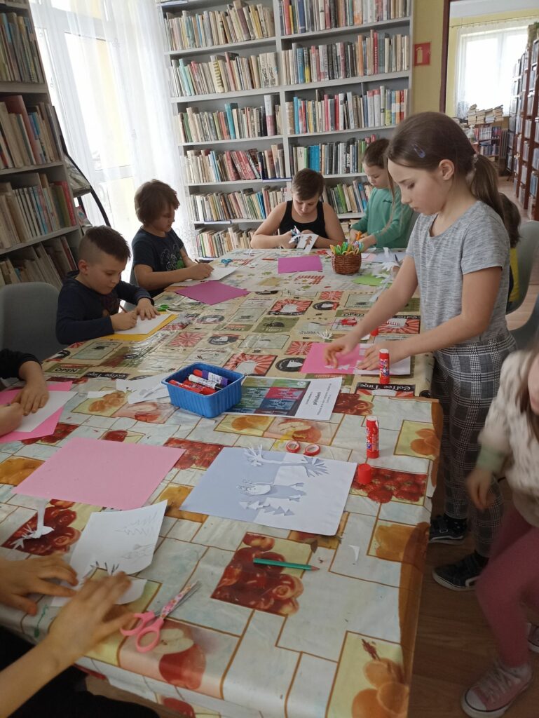 Na zdjęciu widać grupę dzieci siedzących przy stolikach i wykonujących prace plastyczne. Zajęcia odbywają się w bibliotece. W tle regały z książkami.