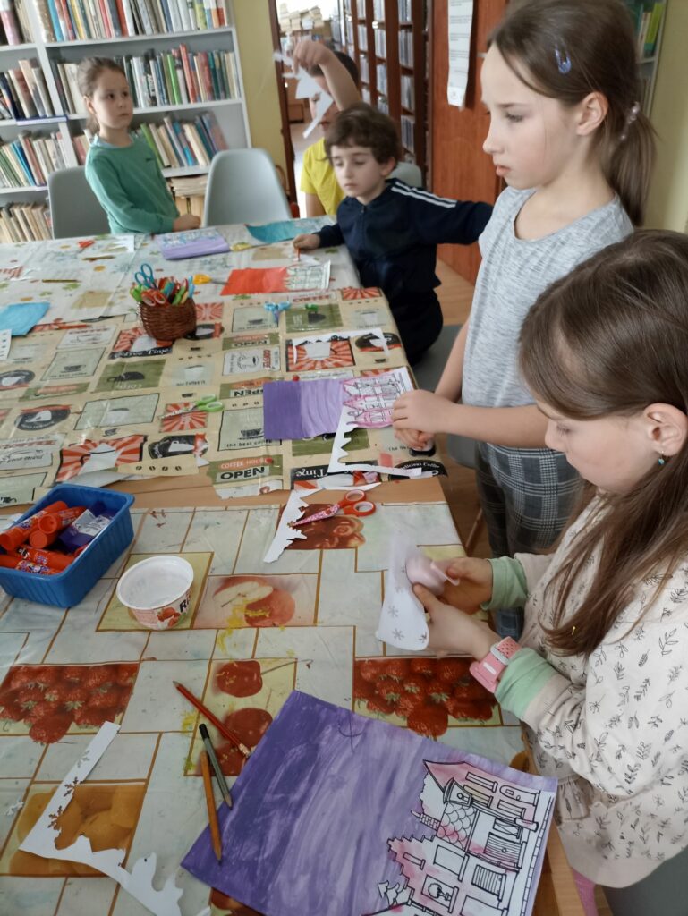 Na zdjęciu widać grupę dzieci siedzących przy stolikach i wykonujących prace plastyczne. Zajęcia odbywają się w bibliotece. W tle regały z książkami.
