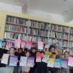 Na zdjęciu widać grupę dzieci (4 chłopców i 5 dziewczynek) pozujących z wykonanymi przez siebie pracami plastycznymi. Dzieci stoją na tle regałów z książkami.