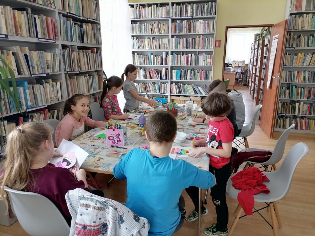 Na zdjęciu widać grupę dzieci (3 chłopców, 5 dziewczynek)siedzących przy stolikach i wykonujących prace plastyczne. Zajęcia odbywają się w bibliotece. W tle regały z książkami.