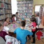Na zdjęciu widać grupę dzieci (3 chłopców, 5 dziewczynek)siedzących przy stolikach i wykonujących prace plastyczne. Zajęcia odbywają się w bibliotece. W tle regały z książkami.