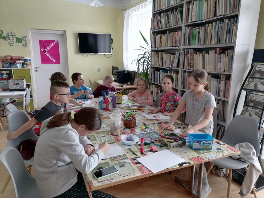 Na zdjęciu widać grupę dzieci (3 chłopców, 5 dziewczynek)siedzących przy stolikach i wykonujących prace plastyczne. Zajęcia odbywają się w bibliotece. W tle regały z książkami, telewizor wiszący na ścianie, kącik gier planszowych.