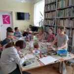 Na zdjęciu widać grupę dzieci (3 chłopców, 5 dziewczynek)siedzących przy stolikach i wykonujących prace plastyczne. Zajęcia odbywają się w bibliotece. W tle regały z książkami, telewizor wiszący na ścianie, kącik gier planszowych.