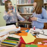 na zdjęciu widać salę biblioteczną, regały z książkami, stół, na stole materiały plastyczne, dwie dziewczynki stoją przy stole