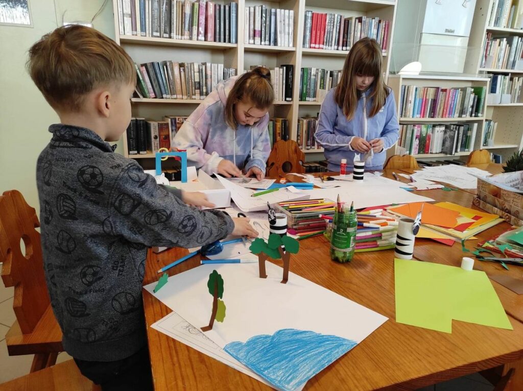 na zdjęciu widać salę biblioteczną, regały z książkami, stół, wokół stołu są dzieci, wykonują pracę plastyczną
