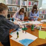 na zdjęciu widać salę biblioteczną, regały z książkami, stół, wokół stołu są dzieci, wykonują pracę plastyczną