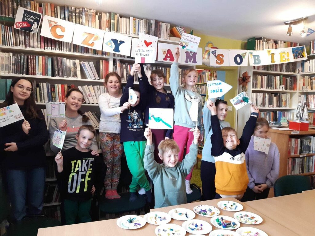 na zdjęciu widać dzieci stojące na tle regału bibliotecznego nad nimi napis czytam sobie