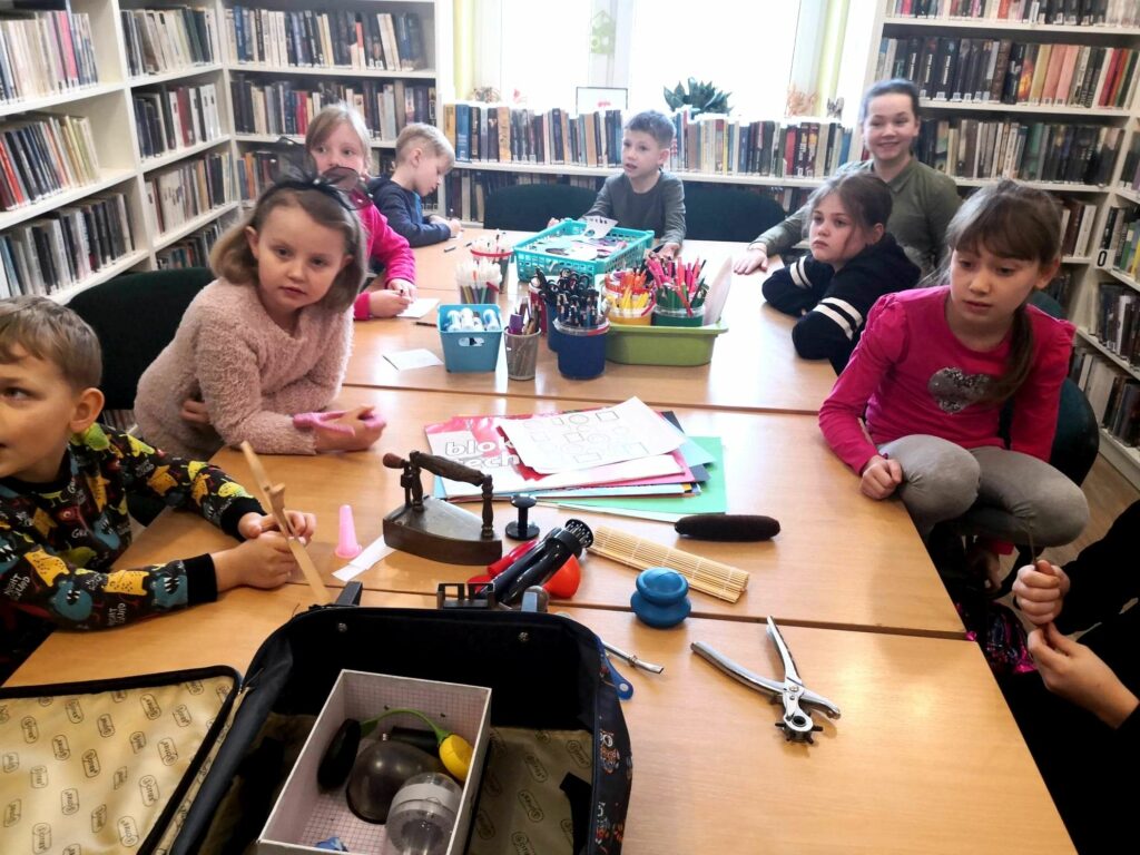 na zdjęciu widać salę biblioteczną, regały z książkami, okno i dzieci siedzące wokół stołu