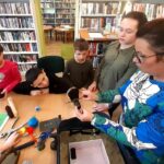 na zdjęciu widać salę biblioteczna siedzące przy stole dzieci i bibliotekarkę pokazującą im różne przedmioty