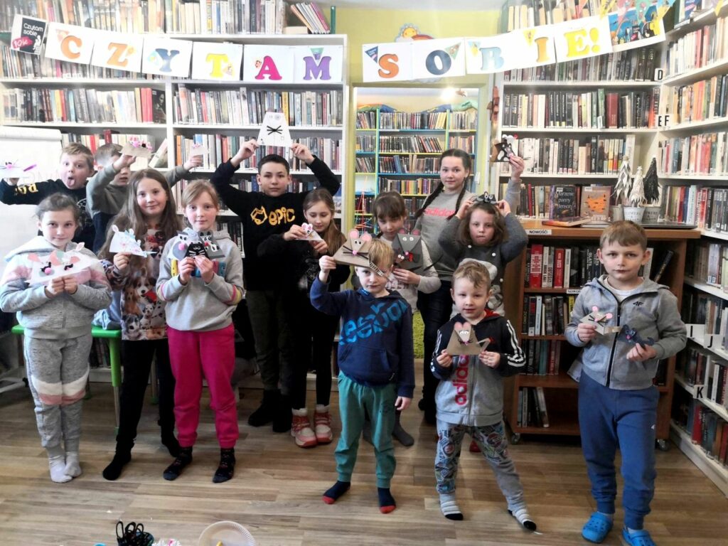 na zdjęciu widać dzieci stojące w sali bibliotecznej na tle regału z książkami