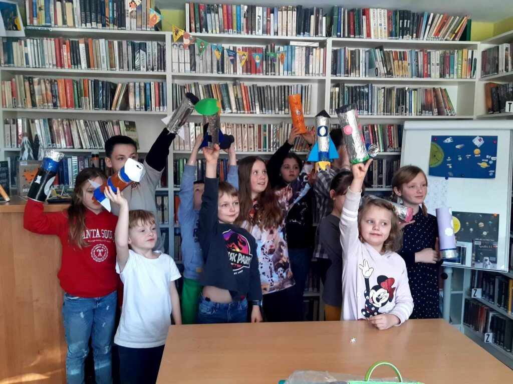 na zdjęciu widać dzieci stojące w sali bibliotecznej na tle regału z książkami