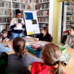 na zdjęciu widać dzieci siedzące wokół stołu w sali bibliotecznej oraz bibliotekarkę pokazującą im zdjęcia