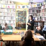 na zdjęciu widać dzieci siedzące wokół stołu w sali bibliotecznej oraz bibliotekarkę czytającą im zagadki, widać półki wypełnione ksiązkanmi