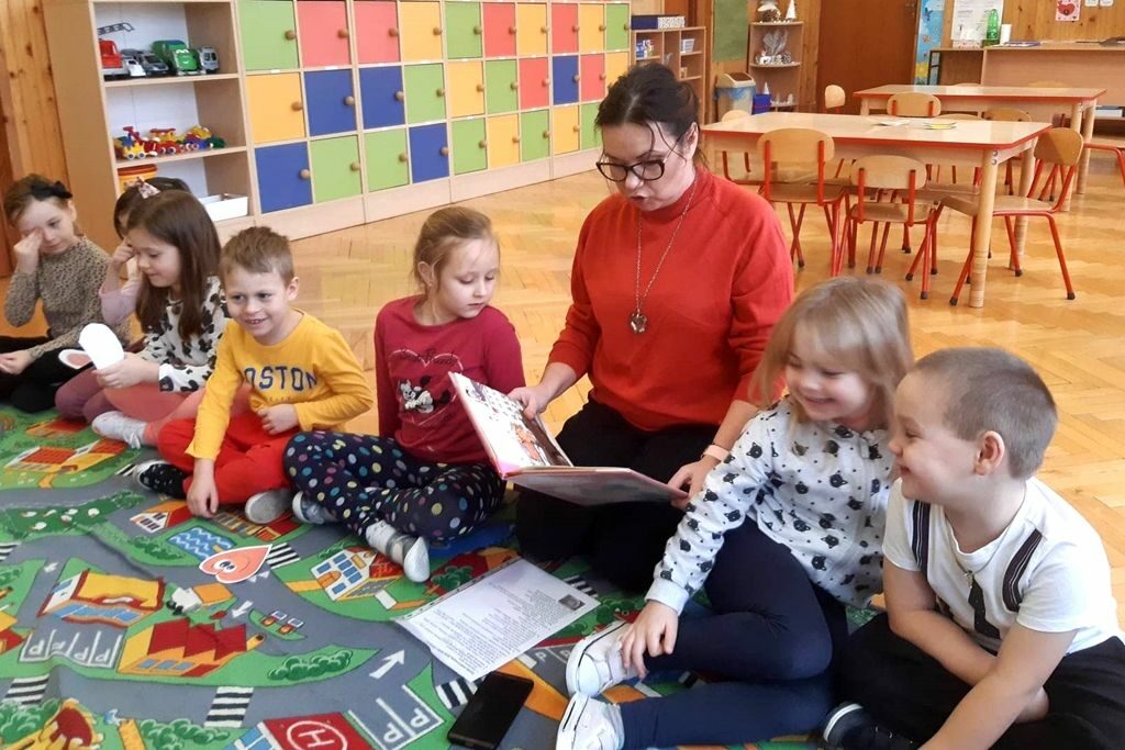 zdjęcie przedstawia bibliotekarkę czytająca dzieciom. Wszyscy siedzą na podłodze