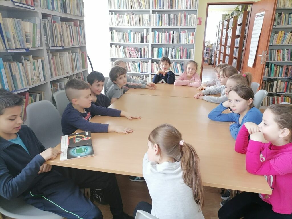 – Dzieci (4 chłopców i 9 dziewczynek) siedzą przy stolikach i czytają na głos książkę pt. „Awantura w śmietniku”. W tle regały z książkami.