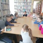 – Dzieci (4 chłopców i 9 dziewczynek) siedzą przy stolikach i czytają na głos książkę pt. „Awantura w śmietniku”. W tle regały z książkami.