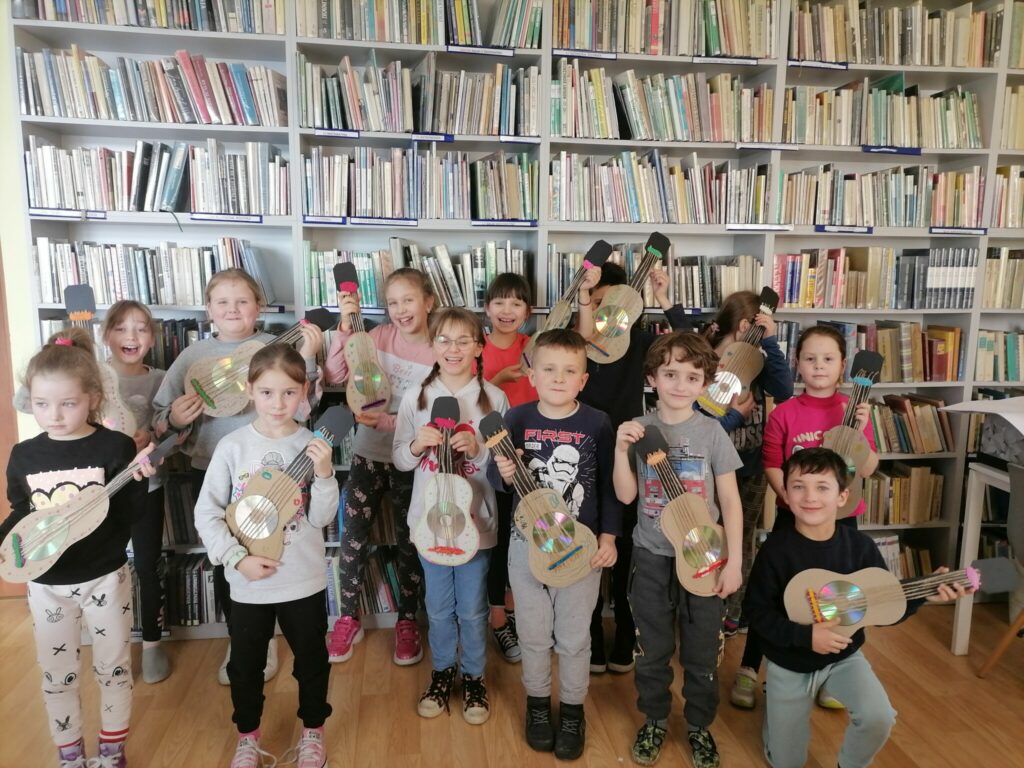 – Dzieci (4 chłopców i 9 dziewczynek) pozują z wykonanymi przez siebie recyklingowymi gitarami, na tle regałów z książkami.