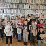 – Dzieci (4 chłopców i 9 dziewczynek) pozują z wykonanymi przez siebie recyklingowymi gitarami, na tle regałów z książkami.
