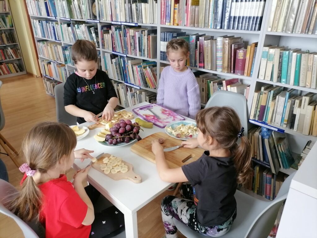 Dzieci (4 dziewczynki) siedzą przy stoliku i kroją owoce (banany, jabłka, winogrona) do sałatki owocowej. W tle regały z książkami.