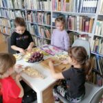 Dzieci (4 dziewczynki) siedzą przy stoliku i kroją owoce (banany, jabłka, winogrona) do sałatki owocowej. W tle regały z książkami.
