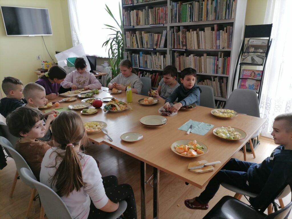 Dzieci (4 dziewczynki i 6 chłopców) siedzą przy stolikach i kroją owoce (kiwi, banany, mandarynki, jabłka, winogrona) do sałatki owocowej. W tle regały z książkami, regalik z gazetami, zawieszony na ścianie telewizor, biurko z komputerami i drukarką oraz duży kwiat.