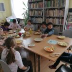 Dzieci (4 dziewczynki i 6 chłopców) siedzą przy stolikach i kroją owoce (kiwi, banany, mandarynki, jabłka, winogrona) do sałatki owocowej. W tle regały z książkami, regalik z gazetami, zawieszony na ścianie telewizor, biurko z komputerami i drukarką oraz duży kwiat.