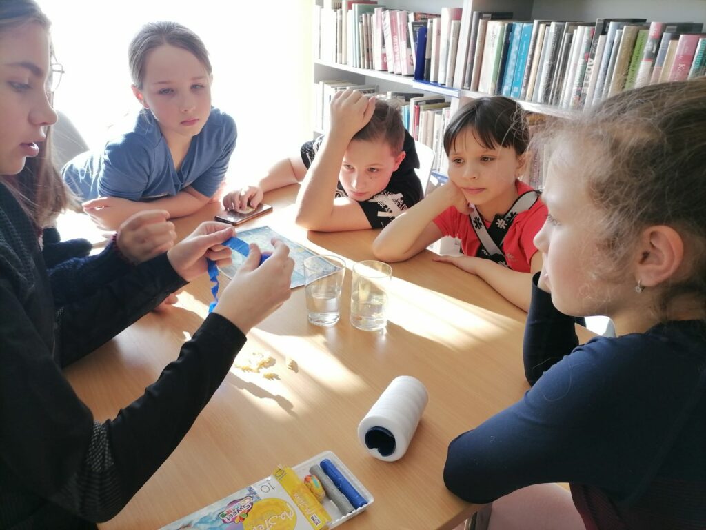 Na zdjęciu widoczne są dzieci (1 chłopiec i 4 dziewczynki) podczas wykonywania eksperymentów wodnych. W tle regały z książkami.