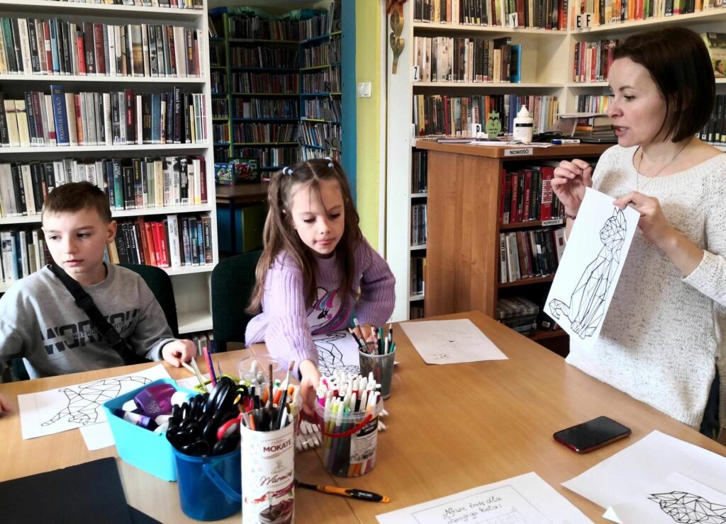 na zdjęciu widać dzieci przy stole oraz bibliotekarkę pokazującą im obrazek