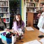 na zdjęciu widać dzieci przy stole oraz bibliotekarkę pokazującą im obrazek