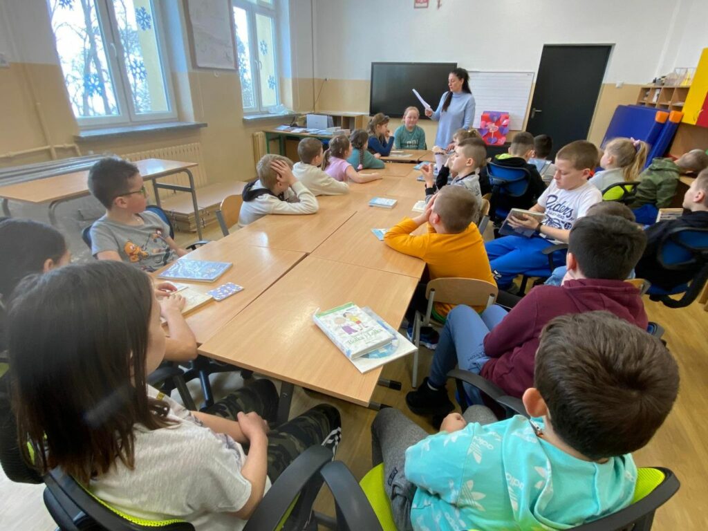 zdjęcie przedstawia salę szkolną, na środku sali stoi stół, wokół stołu siedzą dzieci, bibliotekarz czyta fragment książki