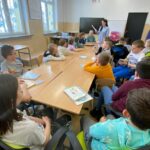 zdjęcie przedstawia salę szkolną, na środku sali stoi stół, wokół stołu siedzą dzieci, bibliotekarz czyta fragment książki