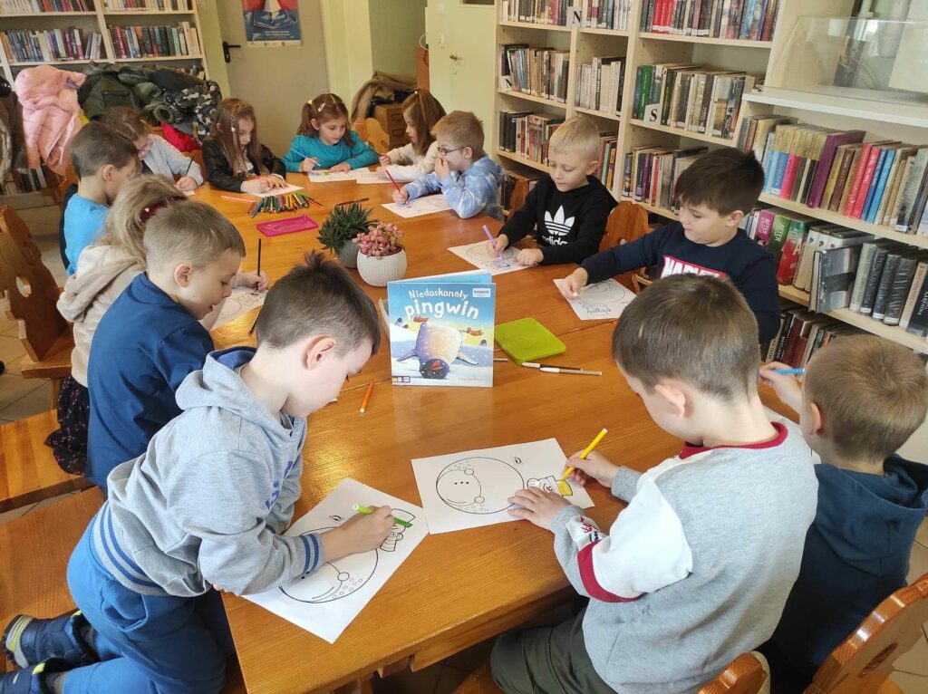 na zdjęciu widać salę biblioteczną, stół, wokół stołu siedzą dzieci, wykonują pracę plastyczną