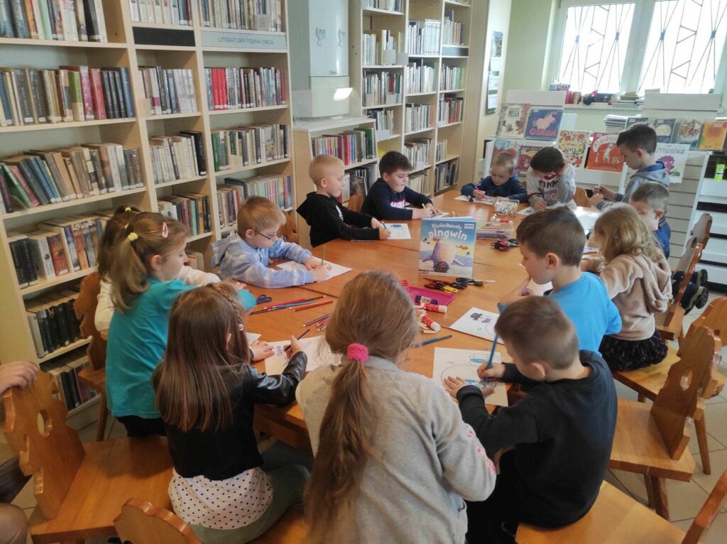 na zdjęciu widać salę biblioteczną, stół, wokół stołu siedzą dzieci, wykonują pracę plastyczną