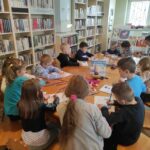 na zdjęciu widać salę biblioteczną, stół, wokół stołu siedzą dzieci, wykonują pracę plastyczną