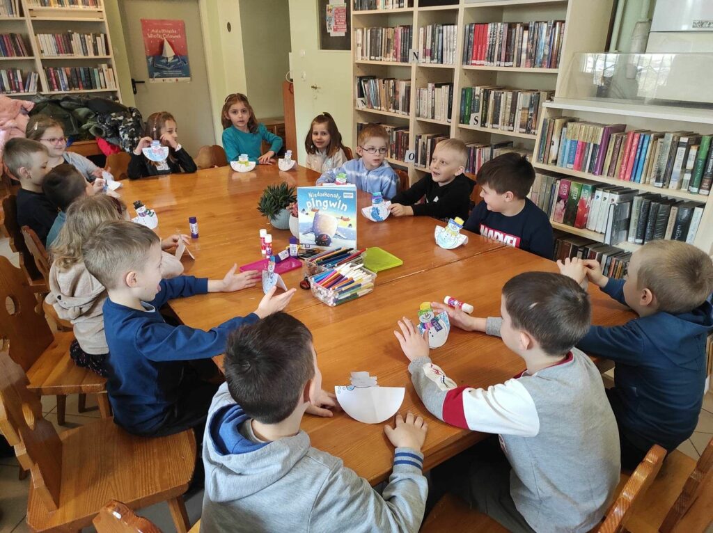 na zdjęciu widać salę biblioteczną, stół, wokół stołu siedzą dzieci, prezentują swoją pracę plastyczną