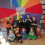 Na zdjęciu widoczna jest grupa przedszkolaków (8 dziewczynek, 3 chłopców) w kolorowych strojach karnawałowych (tygryski, smok, koty, sarenka, motyl, żabka, lew) na tle rozwieszonej na regałach z książkami kolorowej chusty clanzy.