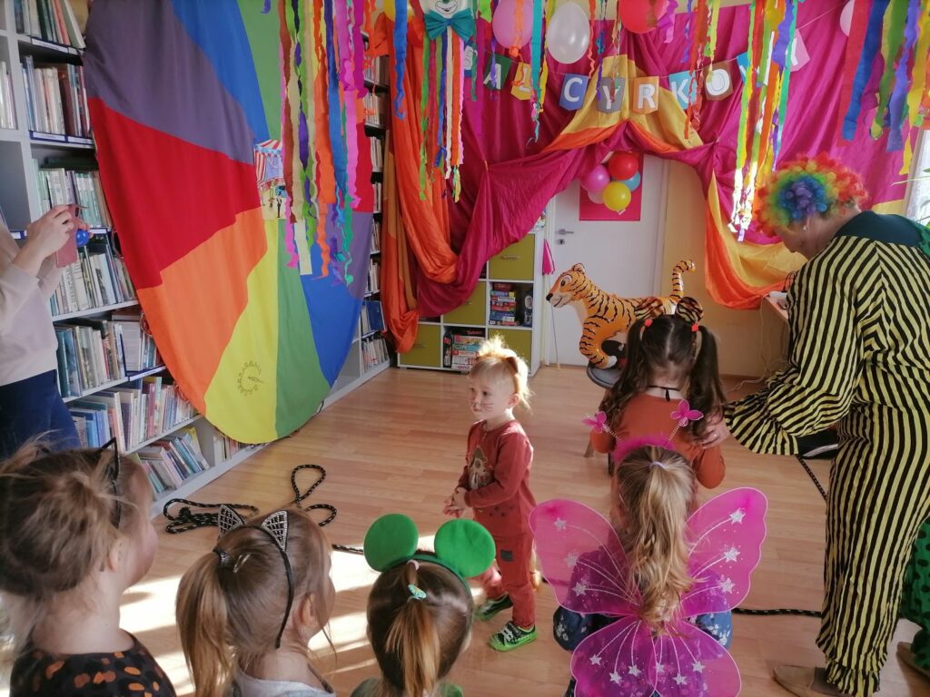 Na zdjęciu widoczne są dzieci oraz bibliotekarz przeprany w strój clowna podczas zabawy w karmienie tygrysa. Dzieci ubrane są w kolorowe stroje. Wokół dekoracje nawiązujące do cyrku.