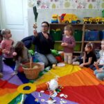 na zdjęciu widać dzieci i bibliotekarkę siedzące na ziemi. Bibliotekarka pokazuje dzieciom por
